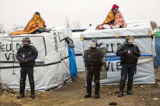 FRANCE, Calais: Riot policer officers stand guard next to migrants on shelters roof as agents dismantle shacks in the Jungle migrant camp, in the French northern port city of Calais, on March 1, 2016. Workers were due to start a second day of destruction in the southern half of the camp, where thousands of migrants and refugees have been living while they try to reach Britain.
