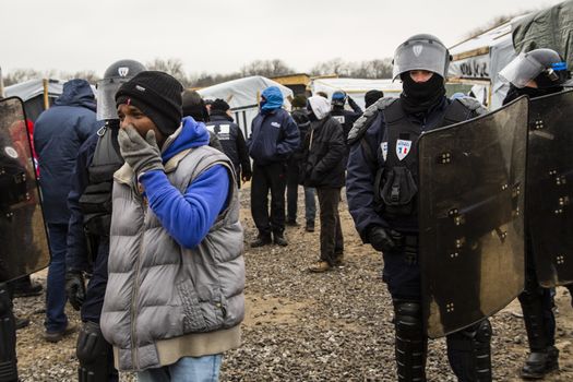 FRANCE, Calais: Riot police officer stand guard next to migrants as agents dismantle shacks and shelters in the Jungle migrant camp, in the French northern port city of Calais, on March 1, 2016. Workers were due to start a second day of destruction in the southern half of the camp, where thousands of migrants and refugees have been living while they try to reach Britain.