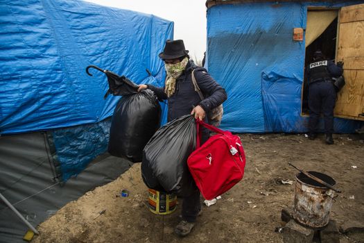 FRANCE, Calais: A migrant leaves his shack, inspected by a police officer, as agents dismantle shelters in the Jungle migrant camp, in the French northern port city of Calais, on March 1, 2016. Workers were due to start a second day of destruction in the southern half of the camp, where thousands of migrants and refugees have been living while they try to reach Britain.