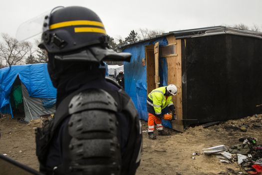 FRANCE, Calais: A police officer stands next to agents dismantling a shack in the Jungle migrant camp, in the French northern port city of Calais, on March 1, 2016. Workers were due to start a second day of destruction in the southern half of the camp, where thousands of migrants and refugees have been living while they try to reach Britain.
