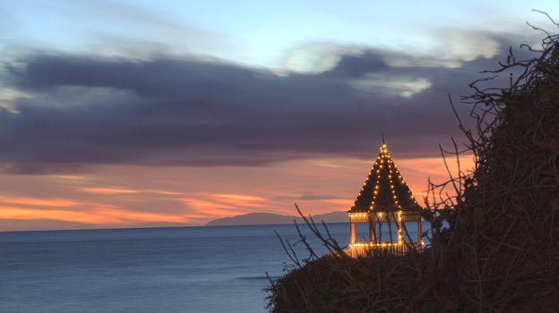 A gazebo on a cliff overlooking the ocean in Laguna Beach, California, lit with Christmas lights at sunset.