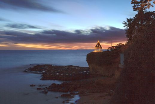 A gazebo on a cliff overlooking the ocean in Laguna Beach, California, lit with Christmas lights at sunset.