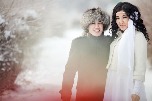 Winter wedding, exotic Asian couple newlyweds in winter attire, groom in fur hat, the bride in winter coats, together looking into the camera. Snowy weather, snow drifts behind.