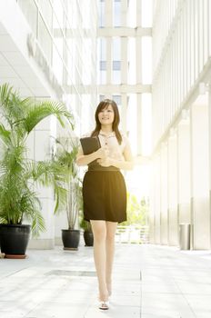 Young Asian business woman smiling and walking at an office building, beautiful golden sunlight at background.