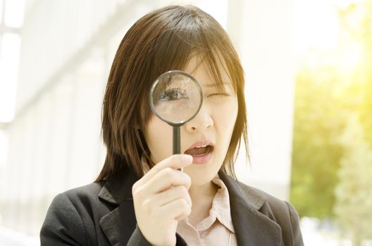 Young Asian business woman looking through magnifier, at an office environment, natural golden sunlight at background.