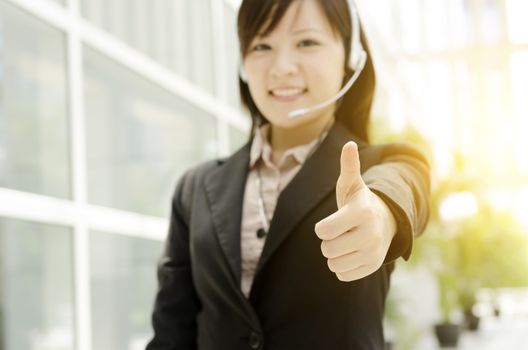 Portrait of a young Asian customer helpline with headset thumb up, at an office environment, natural golden sunlight at background.