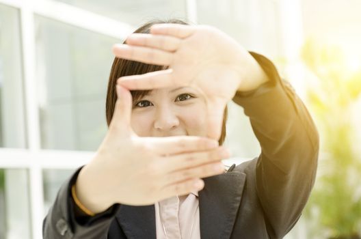 Young Asian business woman creating frame with fingers, at an office environment, natural golden sunlight at background.