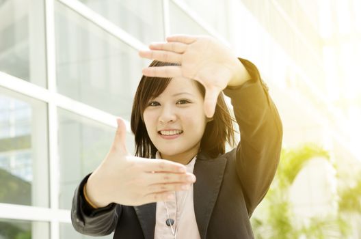 Smiling young Asian business woman making a frame with fingers, at an office environment, natural golden sunlight at background.