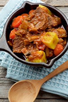 Delicious Beef Stew with Carrots, Potatoes, Celery and Leek in Cast Iron on Blue Napkin with Wooden Spoon closeup. Top View
