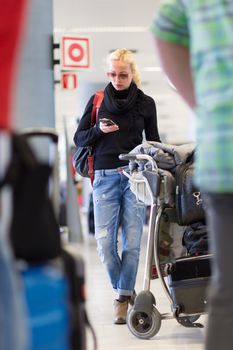 Casual blond young woman using her cell phone while queuing for flight check-in and baggage drop. Wireless network hotspot enabling people to access internet conection. Public transport.