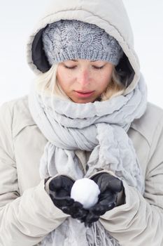 Cute casual young woman wearing glooves, woolen cap and scarf, holding icy snowball in cold winter time. Lady looking down at snowball.