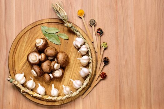 Braid of garlic, mushrooms and bay leaves on a round cutting board light wooden table. Nearby are several small wooden spoons with different spices. Top view.