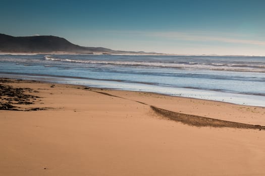 Sandy beach at the Atlantic Ocean shore in Morocco. Small waves of the ocean. Mountain in the background. Early evening little cloudy sky.