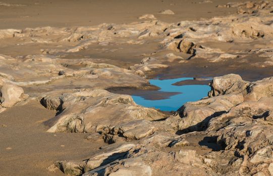 Little rocks in the color of a sand. Puddle in a shape of letter S, reflecting the blue sky. Shore of Atlantic Ocean in Morocco.