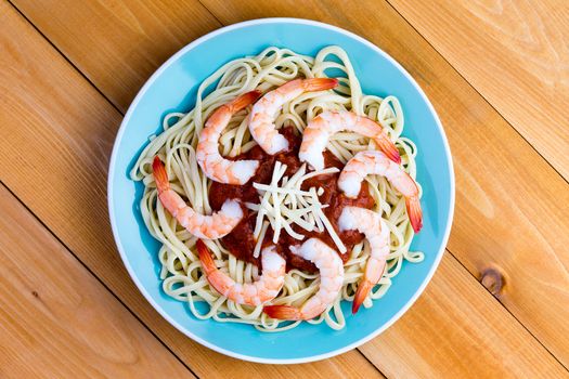 Savory Italian seafood pasta with peeled shrimps on a bed of spaghetti with a tomato sauce and grated gruyere cheese garnish, overhead view on a blue plate on a picnic table