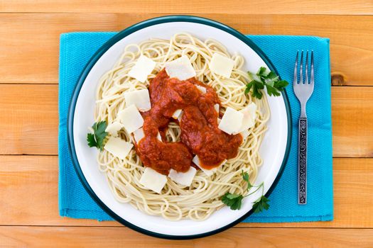 Top down view on plate of spaghetti pasta garnished with grana padano cheese bits, italian parsley and red sauce over blue placemat on wooden table