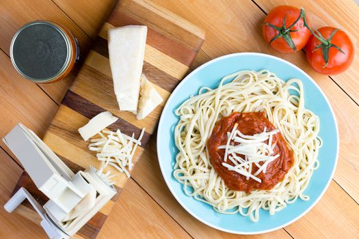 Top down first person perspective view on blue plate of noodles topped with red sauce and grated gruyere cheese next to pasta press and medium tomatoes