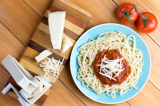 Top down first person perspective view on blue plate of spaghetti topped with red sauce and grated gruyere cheese next to pasta press and two tomatoes