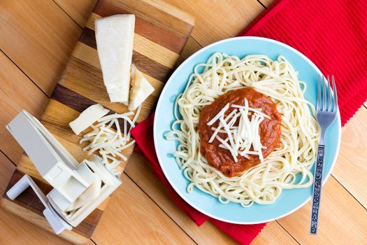 Top down first person perspective view on round plate of freshly cooked spaghetti topped with red sauce and grated gruyere cheese next to pasta press on cutting board