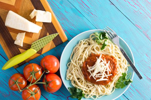 Top down view on round plate of delicious spaghetti topped with red sauce and parsley next to wedge of gruyere cheese and knife on cutting board with vine tomatoes