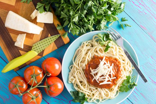 View from above on round plate of freshly prepared spaghetti topped with red sauce, parsley and gruyere cheese beside knife on cutting board with cherry tomatoes