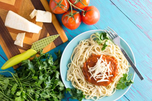 View from above on single plate of freshly prepared spaghetti topped with red sauce, parsley and gruyere cheese next to knife on cutting board with cherry tomatoes over blue table