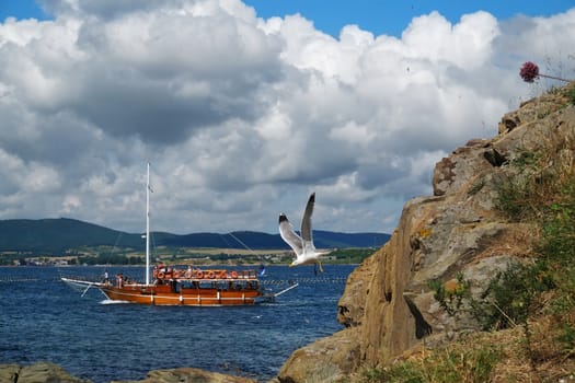 Maritime day landscape with rock, seagull, pleasure boats, sky with clouds and a distant shore. Sozopol, Bulgaria.