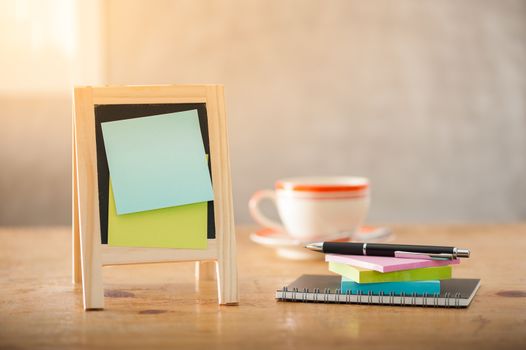 Sticky paper with blank area for text or message stick on small A-frame black board beside pen and notepad on wood table in morning time