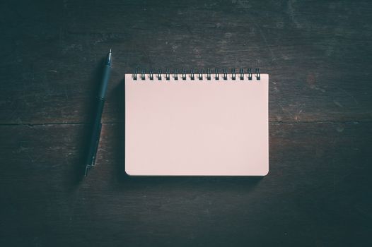 Small opened notepad with blank area for text or message and pen on rustic wood table with low key scene and vinatage filter effect