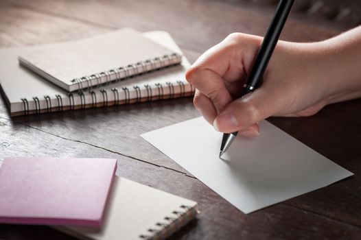 Woman right hand writing on blank paper on wood table with notebooks on wood table with low key scene and vintage filter effect