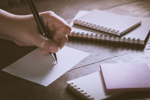Woman left hand writing on blank paper on wood table with notebooks on wood table with low key scene and vintage filter effect