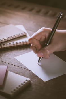 Woman right hand writing on blank paper on wood table with notebooks on wood table with low key scene and vintage filter effect