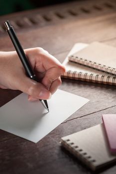 Woman left hand writing on blank paper on wood table with notebooks on wood table with low key scene