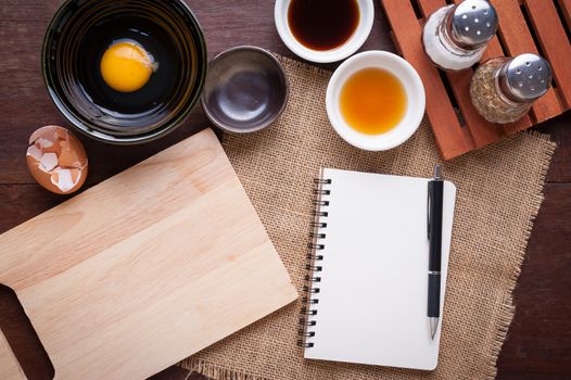 Preparation for cooking in home kitchen. Chopping board, notepad for check list, egg, and ingredients on wood table.