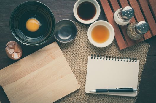 Preparation for cooking in home kitchen. Chopping board, notepad for check list, egg, and ingredients on wood table with vintage filter effect