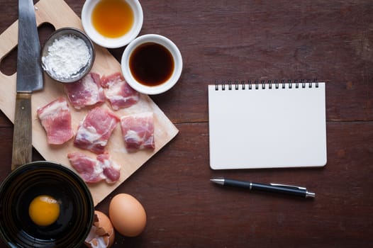 Preparation for cooking in home kitchen. Soft pork ribs on chopping board, notepad for check list, egg, and ingredients on wood table.