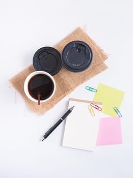 Small notepad with blank space for text or message, sticky paper, pen and take away coffee cup on office table.