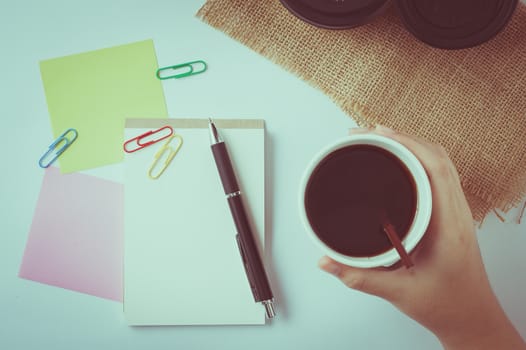 Woman right hand holding take away coffee cup beside notepad on office table with vintage filter effect