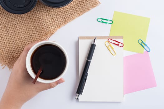 Woman hand holding take away coffee cup beside notepad on office table