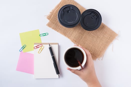 Woman right hand holding take away coffee cup beside notepad on office table
