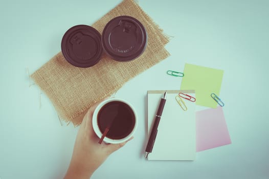 Woman left hand holding take away coffee cup beside notepad on office table with vintage filter effect