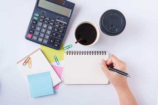 Woman right hand writing on small notepad with blank space for text or message beside sticky paper, calculator, and take away coffee cup on office table