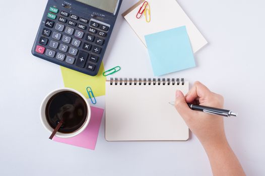 Woman right hand writing on small notepad with blank space for text or message beside sticky paper, calculator, and take away coffee cup on office table