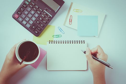 Woman right hand writing on small notepad with blank space for text or message beside sticky paper, calculator, and take away coffee cup on office table while another hand holding coffee cup with vintage filter effect