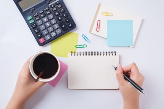 Woman right hand writing on small notepad with blank space for text or message beside sticky paper, calculator, and take away coffee cup on office table while another hand holding coffee cup