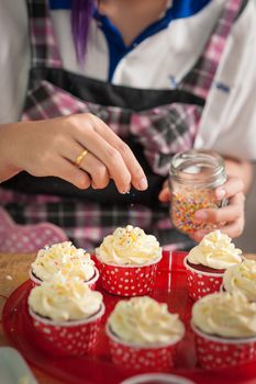 The action of making red velvet cupcakes. Woman hand adding sugar sprinkle on cupcake.