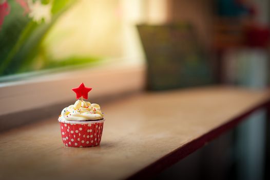 Small red velvet cupcake with cream that made from yogurt and cream cheese garnished with star shape fondant and colourful sugar sprinkle on wood bar in cafe in morning time with low key scene