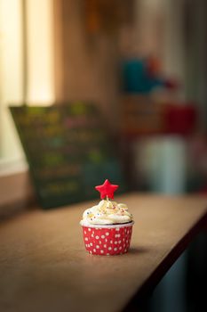 Small red velvet cupcake with cream that made from yogurt and cream cheese garnished with star shape fondant and colourful sugar sprinkle on wood bar in cafe in morning time with low key scene