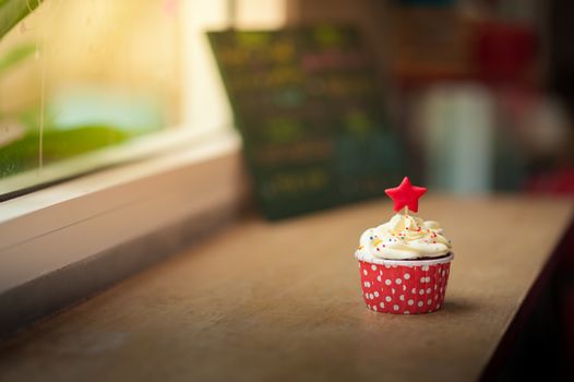Small red velvet cupcake with cream that made from yogurt and cream cheese garnished with star shape fondant and colourful sugar sprinkle on wood bar in cafe in morning time with low key scene