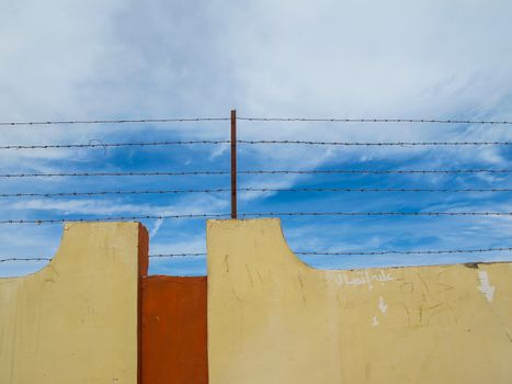 Yellow color of the fence with a barbed wire on the top. Blue sky with intense white clouds.